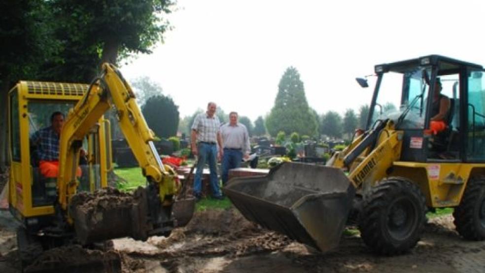 Bagger und Tieflader im Einsatz auf dem Jemgumer Friedhof: Die Wege der von Lindenbäumen umsäumten Anlage hinter der Hofstraße werden saniert. Die Kirchenratsmitglieder Reiner Middelborg (Mitte, links) und Bernhard Spekker begutachten die Arbeiten. © Foto: Hanken