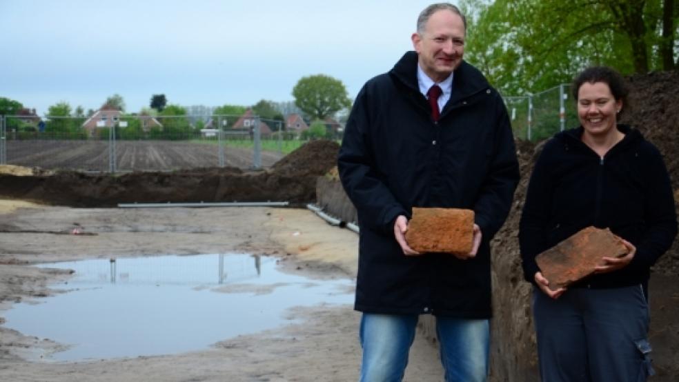 Zwei dicke Brocken Geschichte halten Jonas Werner (Niedersächsische Landgesellschaft) und Archäologin Kirsten Hüser mit zwei Klosterformat-Steinen in den Händen. Dunkel verfärbt zeichnet sich im Hintergrund der Graben des wohl sechsten von sieben Steinhäusern in Stapelmoor ab. © Foto: Wübbena