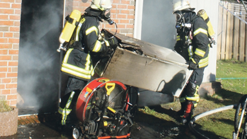 Feuerwehrmänner tragen einen verkohlten Gefrierschrank ins Freie. Die Ursache für den Brand in Tichelwarf ist noch unklar. © Foto: Rand (Feuerwehr)