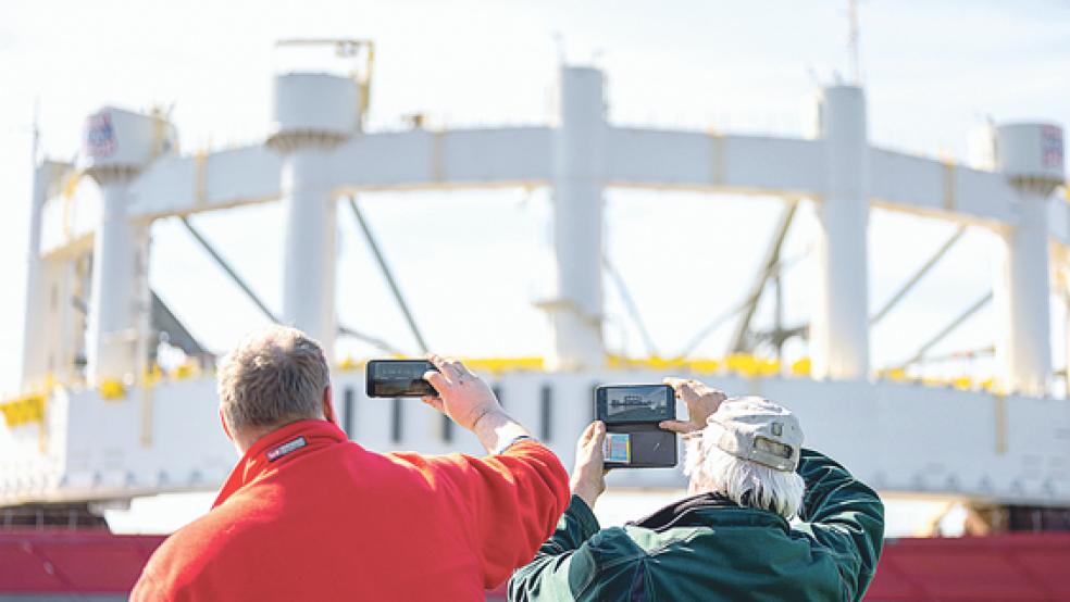 Zwei Männer fotografieren, wie das 1600 Tonnen schwere Bauteil für eine Hochsee-Lachsfarm auf einem Ponton aus dem Emden Hafen bewegt wird.  © Foto: Assanimoghaddam (dpa)