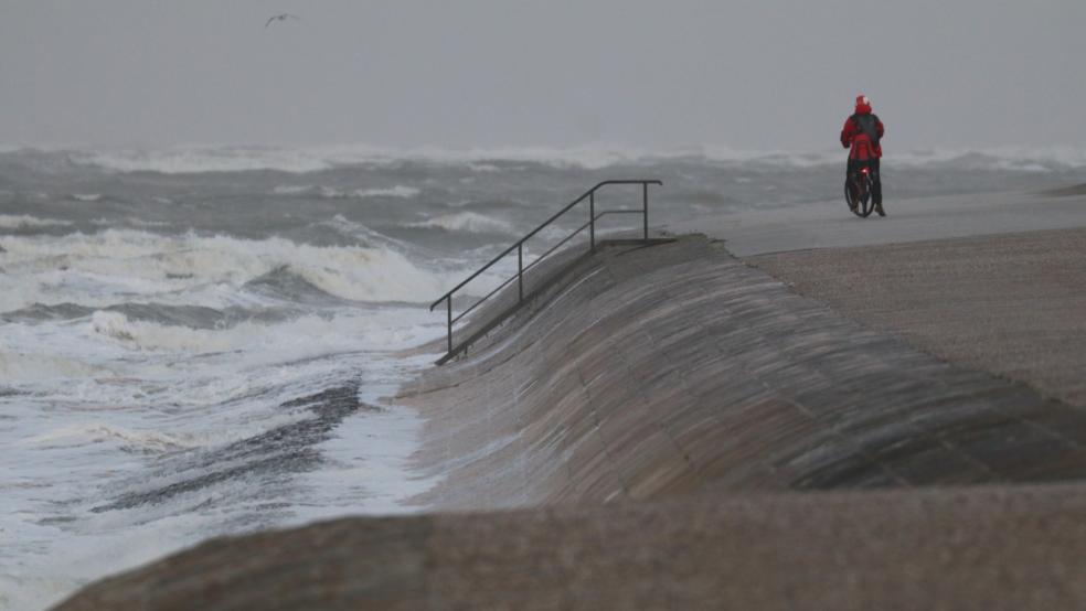 Eine Radfahrerin steht bei stürmischem Wetter an der Wasserkante auf der Insel Norderney.  © dpa/Volker Bartels