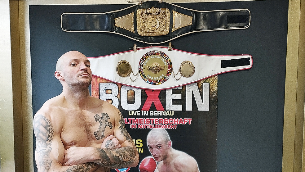 Chris Herrmann präsentiert sich in seinem Tattoostudio in Weener vor seiner Boxwand, die seine beiden Weltmeistergürtel und das Kampfplakat zum dritten Titelkampf 2014 zeigt.  © Foto: Born